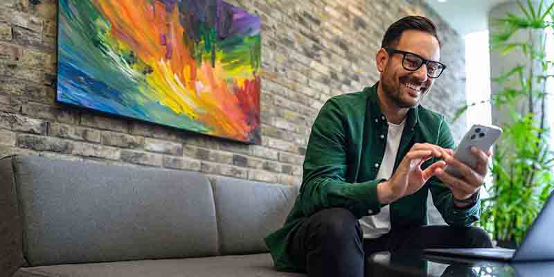 Man in a green shirt looking at his phone while sitting on a couch with a colorful oil painting on the wall behind him.